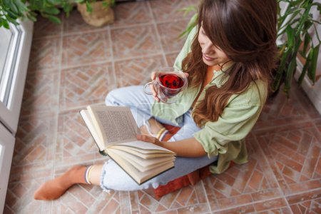 Téléchargez les photos : Vue en angle élevé de la belle jeune femme assise à son balcon, buvant du thé et lisant un livre tout en se relaxant à la maison. Focus sur la tasse - en image libre de droit
