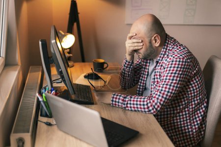 Photo for Web developer sitting at his desk while working in home office, holding head in hands, stressed out because of failure and code malfunction - Royalty Free Image