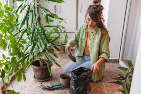Téléchargez les photos : Femme relaxante à la maison en jardinant, plantant des plantes de maison dans des pots de fleurs, ajoutant du sol à l'aide d'une petite pelle - en image libre de droit