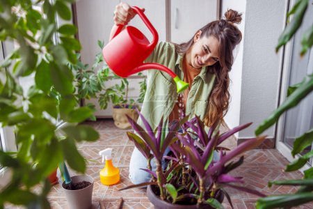 Téléchargez les photos : Belle jeune femme relaxante à la maison, tenant un seau, arrosant et prenant soin de plantes d'intérieur sur son balcon - en image libre de droit
