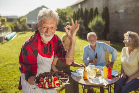 Téléchargez les photos : Homme âgé apportant de la nourriture délicieuse sur un plateau tout en prenant un déjeuner en plein air avec des amis dans la cour de sa maison, montrant un signe OK et souriant - en image libre de droit
