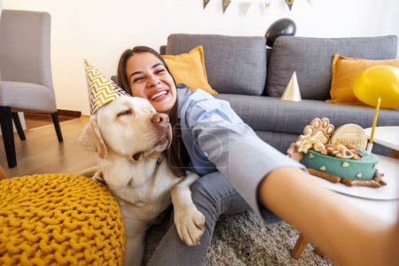Foto de Hermosa joven alegre divirtiéndose tomando selfies mientras celebra el cumpleaños de su perro labrador mascota en casa - Imagen libre de derechos