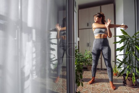 Téléchargez les photos : Jeune femme en vêtements de sport faisant de l'exercice à la maison le matin, étirant les bras après l'entraînement - en image libre de droit