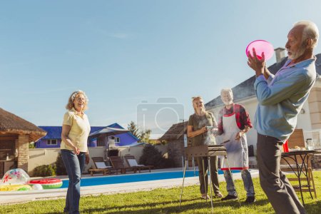 Photo for Group of cheerful active senior people having a backyard barbecue party, grilling meat and vegetables and having fun playing toss and catch - Royalty Free Image
