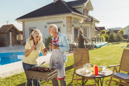 Téléchargez les photos : Groupe de personnes âgées actives gaies ayant une fête barbecue arrière-cour, griller de la viande et des légumes et avoir du plaisir à jouer au badminton - en image libre de droit