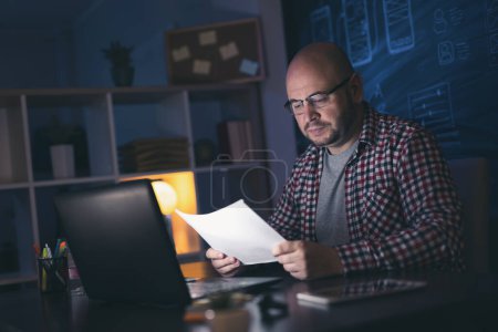 Téléchargez les photos : Homme assis à son bureau dans son bureau à la maison tard dans la nuit, faisant de la paperasse - en image libre de droit