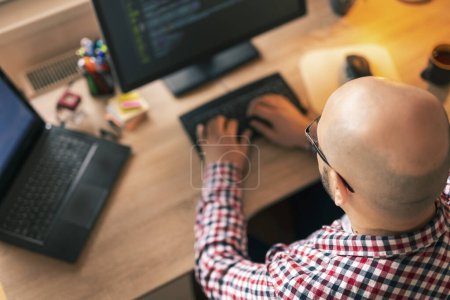 Photo for High angle view of software developer working in home office, sitting at his desk and coding; programmer working remotely from home - Royalty Free Image