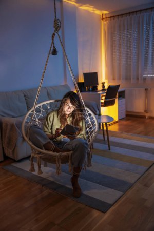 Téléchargez les photos : Belle jeune femme assise dans une chaise suspendue surfer sur le net à l'aide d'un ordinateur tablette tout en se relaxant à la maison - en image libre de droit