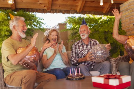 Foto de Grupo de alegres amigos mayores divirtiéndose en una fiesta de cumpleaños, usando sombreros de fiesta, sosteniendo globos y aplaudiendo mientras cantan canción de cumpleaños - Imagen libre de derechos
