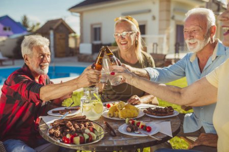 Photo for Group of cheerful senior friends making a toast while having an outdoor lunch in the backyard by the pool, gathered around the table, having fun spending time together - Royalty Free Image