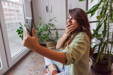 Téléchargez les photos : Belle jeune femme jouissant de loisirs à la maison, ayant un appel vidéo à l'aide d'un téléphone intelligent tout en se relaxant sur son balcon - en image libre de droit