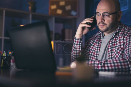 Photo for Man sitting at his desk in home office, having a phone conversation using smart phone while working late at night - Royalty Free Image