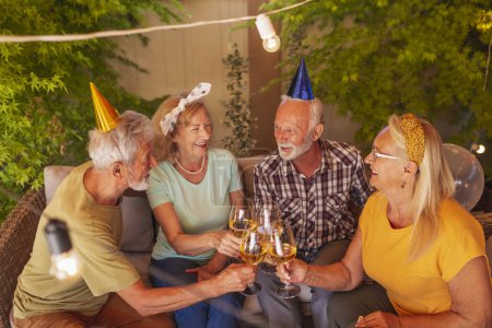 Photo for Group of active cheerful senior friends wearing party hats having fun celebrating birthday, making a toast and drinking wine - Royalty Free Image
