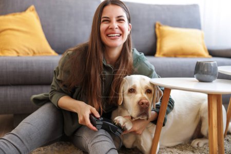 Téléchargez les photos : Jeune femme s'amusant passer du temps libre à la maison avec son animal de compagnie, beau chien du Labrador, jouer à des jeux vidéo et manger du maïs soufflé - en image libre de droit