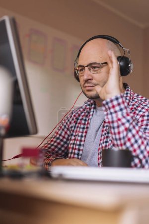 Photo for Man wearing headset and sitting at his desk in home office, having online meeting while working remotely from home - Royalty Free Image