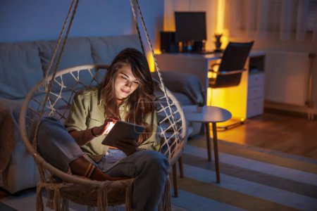 Foto de Hermosa joven sentada en silla colgante leyendo un libro electrónico usando ereader mientras se relaja en casa - Imagen libre de derechos