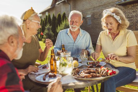 Foto de Grupo de alegres amigos mayores almorzando al aire libre en el patio trasero, reunidos alrededor de la mesa, comiendo, bebiendo y teniendo una agradable conversación - Imagen libre de derechos