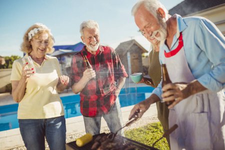 Téléchargez les photos : Groupe d'amis seniors faisant une fête barbecue dans la cour près de la piscine, réunis autour du gril, griller de la viande et s'amuser - en image libre de droit