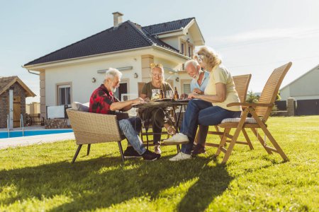Foto de Grupo de alegres amigos mayores almorzando al aire libre en el patio trasero junto a la piscina, reunidos alrededor de la mesa, comiendo, bebiendo y teniendo una agradable conversación - Imagen libre de derechos