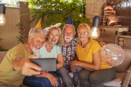 Photo for Group of cheerful senior people having fun celebrating friend's birthday, wearing party hats and taking selfies using smart phone - Royalty Free Image
