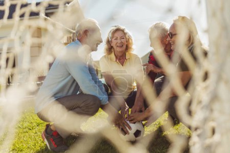 Photo for Group of senior people squatting in front of a goal, gathered in circle, holding a soccer ball all together before playing football in the backyard - Royalty Free Image