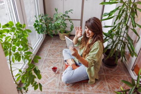 Téléchargez les photos : Vue en angle élevé de la femme passant du temps libre à se détendre à la maison, assis sur son balcon à boire du thé et d'avoir un appel vidéo avec un ami à l'aide de la tablette - en image libre de droit