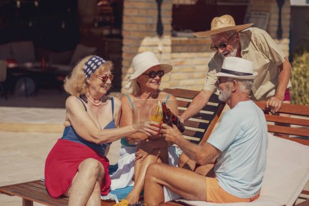 Photo for Group of senior friends relaxing and sunbathing on sun beds by the swimming pool while on a summer vacation, making a toast, drinking cocktails and beer and having fun - Royalty Free Image