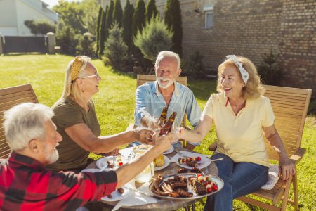Photo for Group of senior neighbors making a toast while having an outdoor lunch in the backyard, gathered around the table, having fun spending time together - Royalty Free Image
