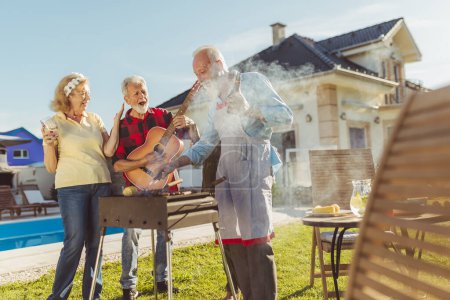 Photo for Group of cheerful elderly people having a poolside backyard barbecue party, gathered around the grill, playing the guitar, singing, grilling meat and having fun - Royalty Free Image