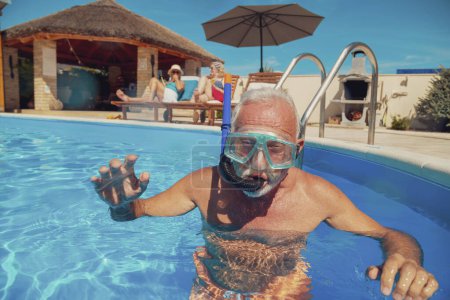 Foto de Grupo de amigos mayores relajándose y disfrutando de las vacaciones de verano, hombre mayor divirtiéndose haciendo snorkel en la piscina mientras las mujeres toman el sol en las tumbonas junto a la piscina - Imagen libre de derechos