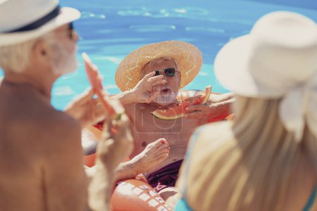 Foto de Grupo de personas mayores alegres sentados en el borde de una piscina comiendo sandía y divirtiéndose al aire libre en un día caluroso de verano - Imagen libre de derechos