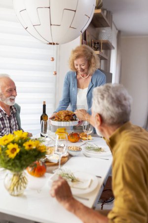 Photo for Two senior couples gathered around table, celebrating Thanksgiving, having dinner together at home, hostess welcoming guests and serving roasted chicken - Royalty Free Image