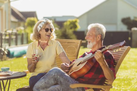 Photo for Cheerful senior couple having an outdoor lunch in the backyard, sitting at the table, drinking beer and lemonade, playing the guitar, singing and relaxing on a sunny summer day - Royalty Free Image