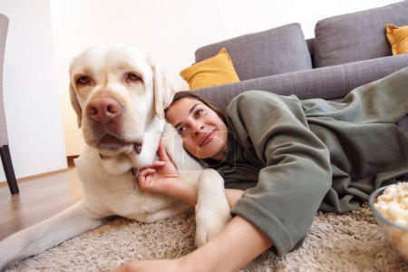 Téléchargez les photos : Belle jeune femme câlinant son chien de compagnie tout en se relaxant et en profitant de loisirs à la maison, allongé sur le sol du salon en mangeant du pop-corn et en regardant la télévision - en image libre de droit