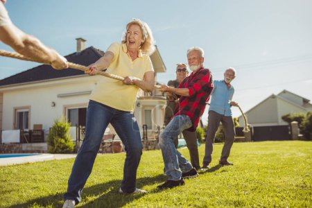 Photo for Senior people having fun playing tug of war game, spending sunny summer day outdoors; group of elderly friends having fun participating in rope pulling competition - Royalty Free Image
