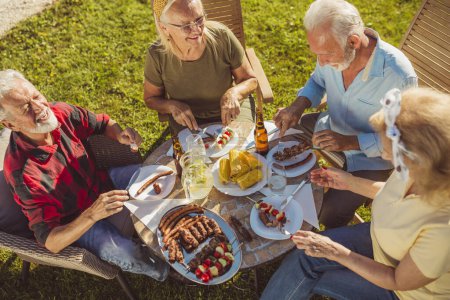 Téléchargez les photos : Vue en angle élevé du groupe d'amis âgés qui déjeunent en plein air dans la cour arrière, réunis autour de la table, mangeant, buvant et s'amusant - en image libre de droit