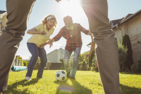 Foto de Grupo de personas mayores activas que se divierten jugando al fútbol en el césped en el patio trasero, disfrutando de un soleado día de verano al aire libre, corriendo tras una pelota - Imagen libre de derechos