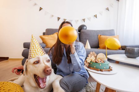 Foto de Joven mujer divirtiéndose soplando globos para su mascota labrador perro fiesta de cumpleaños - Imagen libre de derechos
