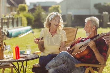 Foto de Alegre pareja mayor teniendo un almuerzo al aire libre en el patio trasero, sentado a la mesa, bebiendo cerveza y limonada, tocando la guitarra, cantando y relajándose en un soleado día de verano - Imagen libre de derechos