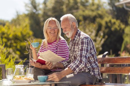 Photo for Happy senior couple enjoying their time together having an outdoor breakfast in the backyard of their home, man reading newspaper while woman is reading a book - Royalty Free Image