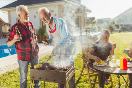 Foto de Grupo de personas mayores divirtiéndose en la fiesta de barbacoa en el patio trasero, hombres asando carne y bebiendo cerveza mientras las mujeres están sentadas y relajándose en el fondo - Imagen libre de derechos