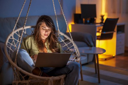 Photo for Woman enjoying leisure time at home, sitting in hanging chair in living room, holding laptop computer in her lap, working remotely from home late at night - Royalty Free Image