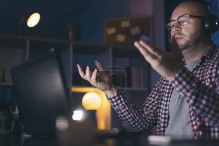 Foto de Hombre freelancer que trabaja hasta tarde desde casa sentado en su escritorio, teniendo una reunión en línea con un cliente usando un ordenador portátil - Imagen libre de derechos