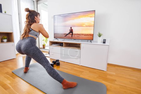 Photo for Active young woman doing online yoga session at home, following exercises shown by instructor on TV - Royalty Free Image