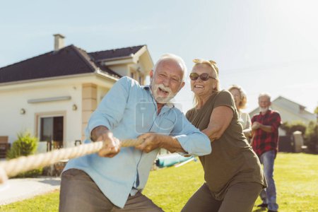 Photo for Senior people having fun playing tug of war game, spending sunny summer day outdoors; group of elderly friends having fun participating in rope pulling competition - Royalty Free Image