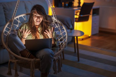 Photo for Woman enjoying leisure time at home, sitting in hanging chair in living room, having video call using laptop computer - Royalty Free Image