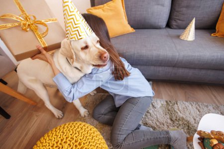 Téléchargez les photos : Belle jeune femme s'amuse à faire une fête d'anniversaire pour son chien à la maison, à l'embrasser et à lui offrir un cadeau d'anniversaire - en image libre de droit