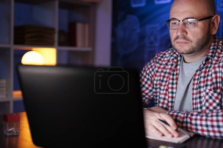 Téléchargez les photos : Homme assis à son bureau, utilisant un ordinateur portable prenant un cours en ligne tard dans la nuit - en image libre de droit