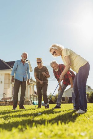 Téléchargez les photos : Groupe d'amis seniors s'amuser à jouer au mini-golf sur la pelouse arrière, passer une journée ensoleillée en plein air - en image libre de droit