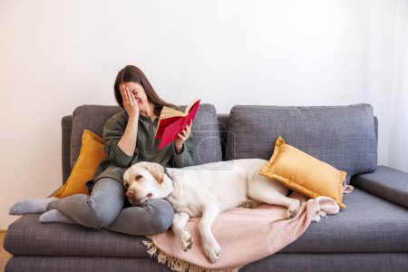 Téléchargez les photos : Belle jeune femme assise sur le canapé du salon lisant un livre tandis que son chien de compagnie est couché à côté d'elle avec la tête sur ses genoux - en image libre de droit